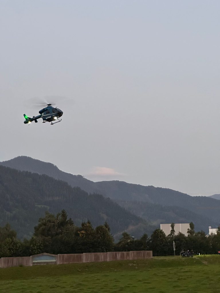 Polizeihubschrauber fliegt über Wiese vor bewaldeten Bergen bei Abenddämmerung