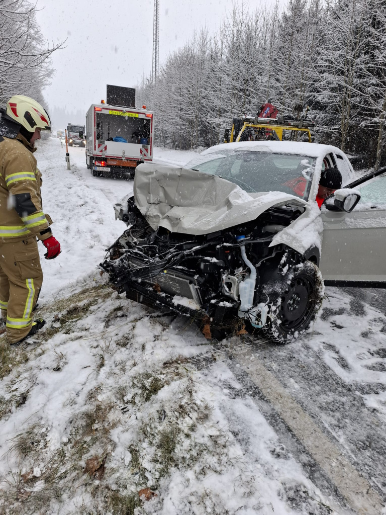 stark beschädigter PKW im Schnee mit Feuerwehrkraft und Einsatzfahrzeugen im Hintergrund