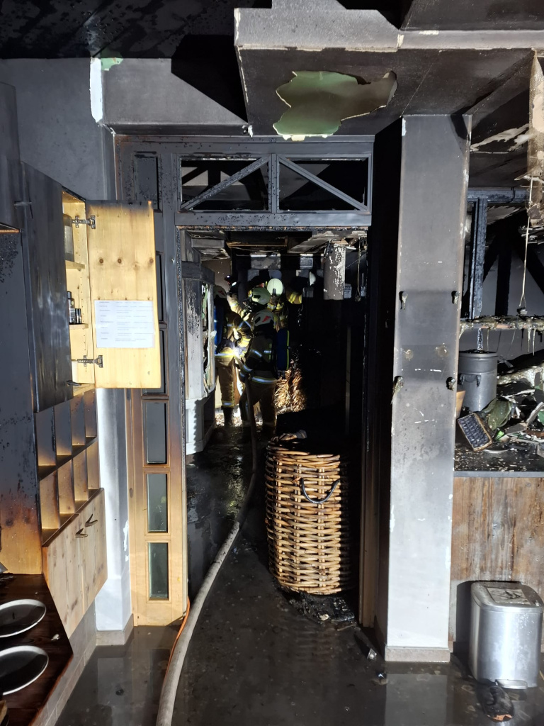 Feuerwehrkräfte in einem verbrannten Hotelflur mit Ruß, Wasserrückständen und beschädigter Decke