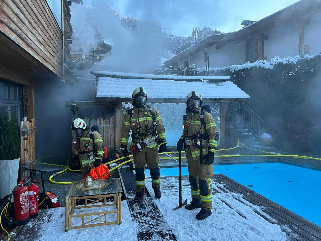 Feuerwehrkräfte in Schutzanzügen arbeiten vor einem rauchenden Wellnessbereich neben einem verschneiten Außenpool